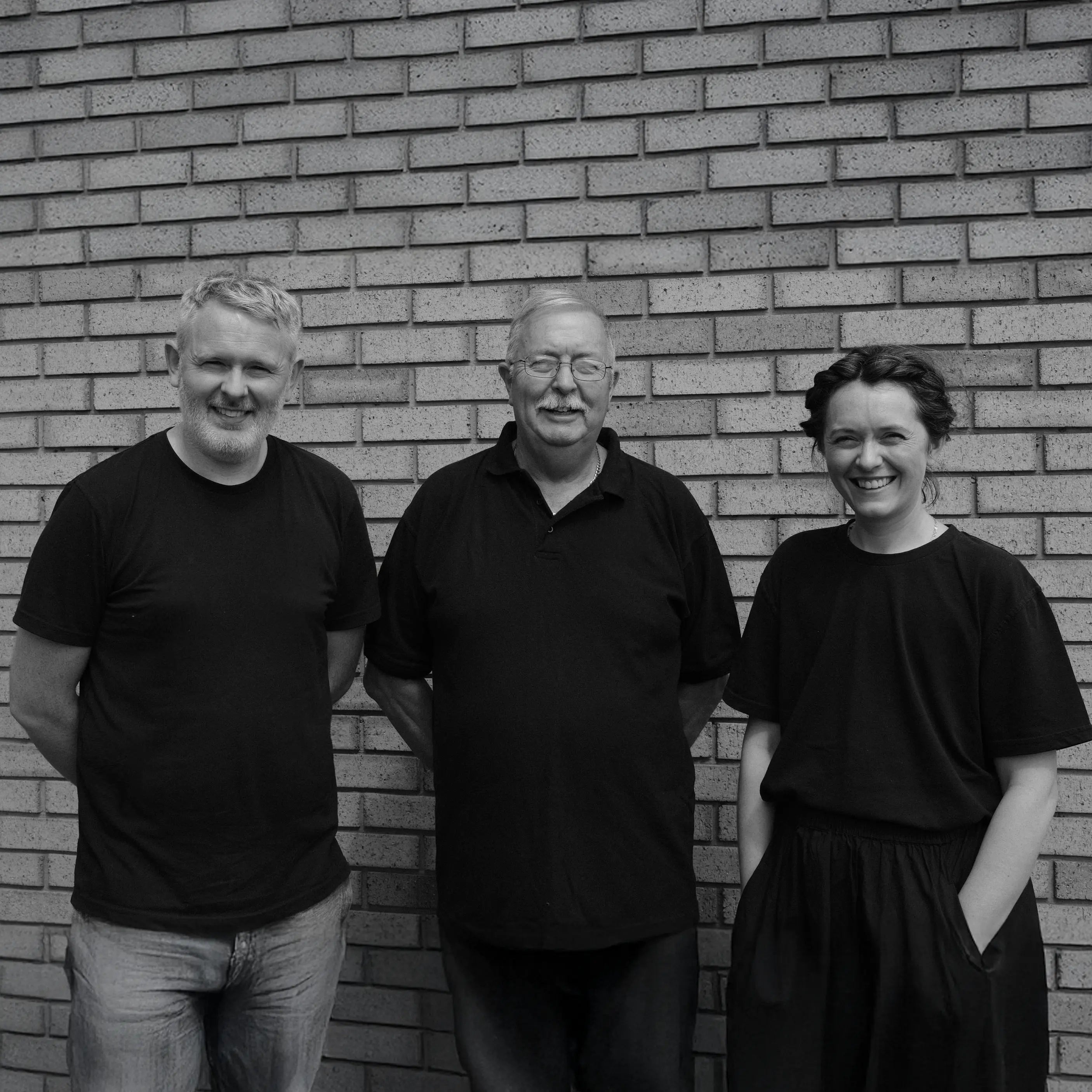 A black-and-white photograph of three people standing side by side against a tiled brick wall. On the left is a middle-aged man with short, light-colored hair and a short beard, wearing a dark T-shirt and light-colored pants. In the center is an older gentleman with glasses, a mustache, and short hair, dressed in a collared shirt and dark pants. On the right is a smiling young woman with her hair braided and pulled back, wearing a dark T-shirt